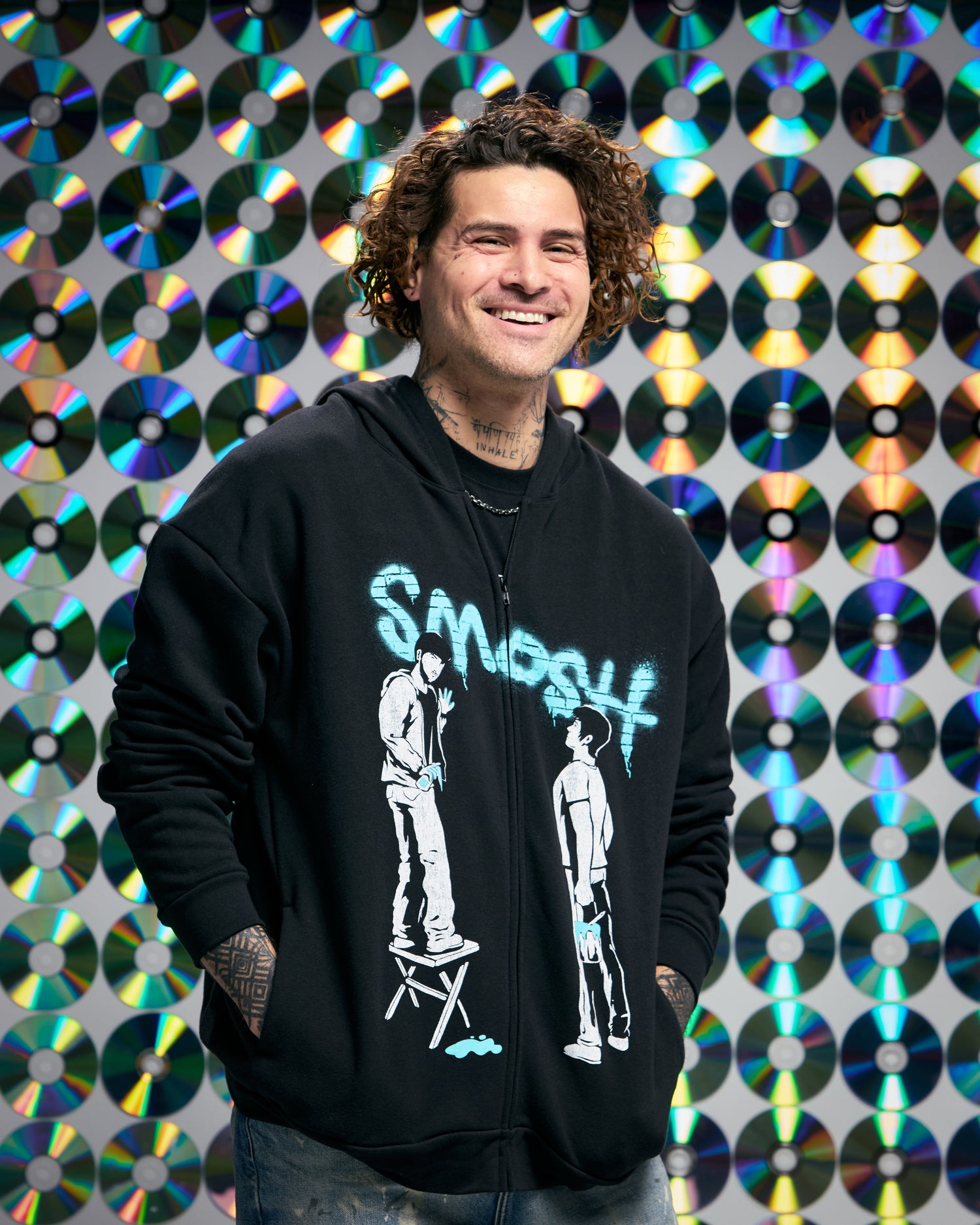 Person wearing a black hoodie with graphic design in front of a colorful CD wall.