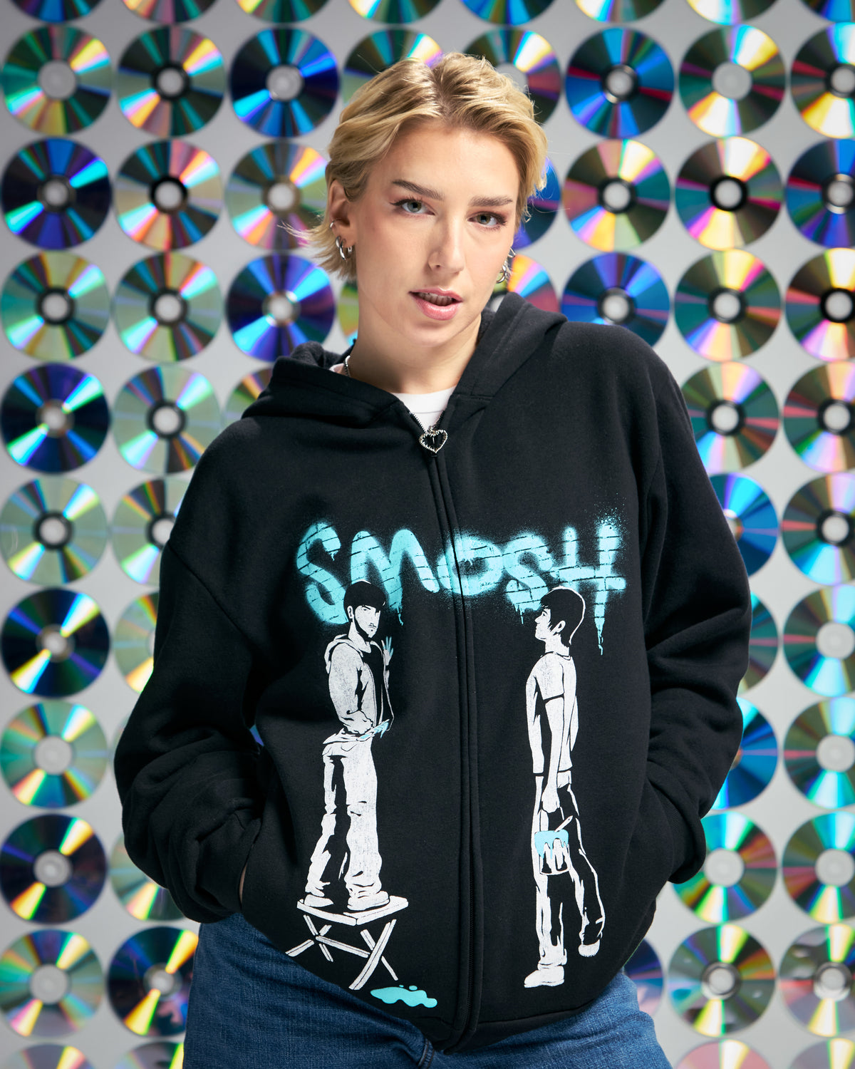Person wearing a black hoodie with Smosh design and text, standing in front of a colorful CD wall.