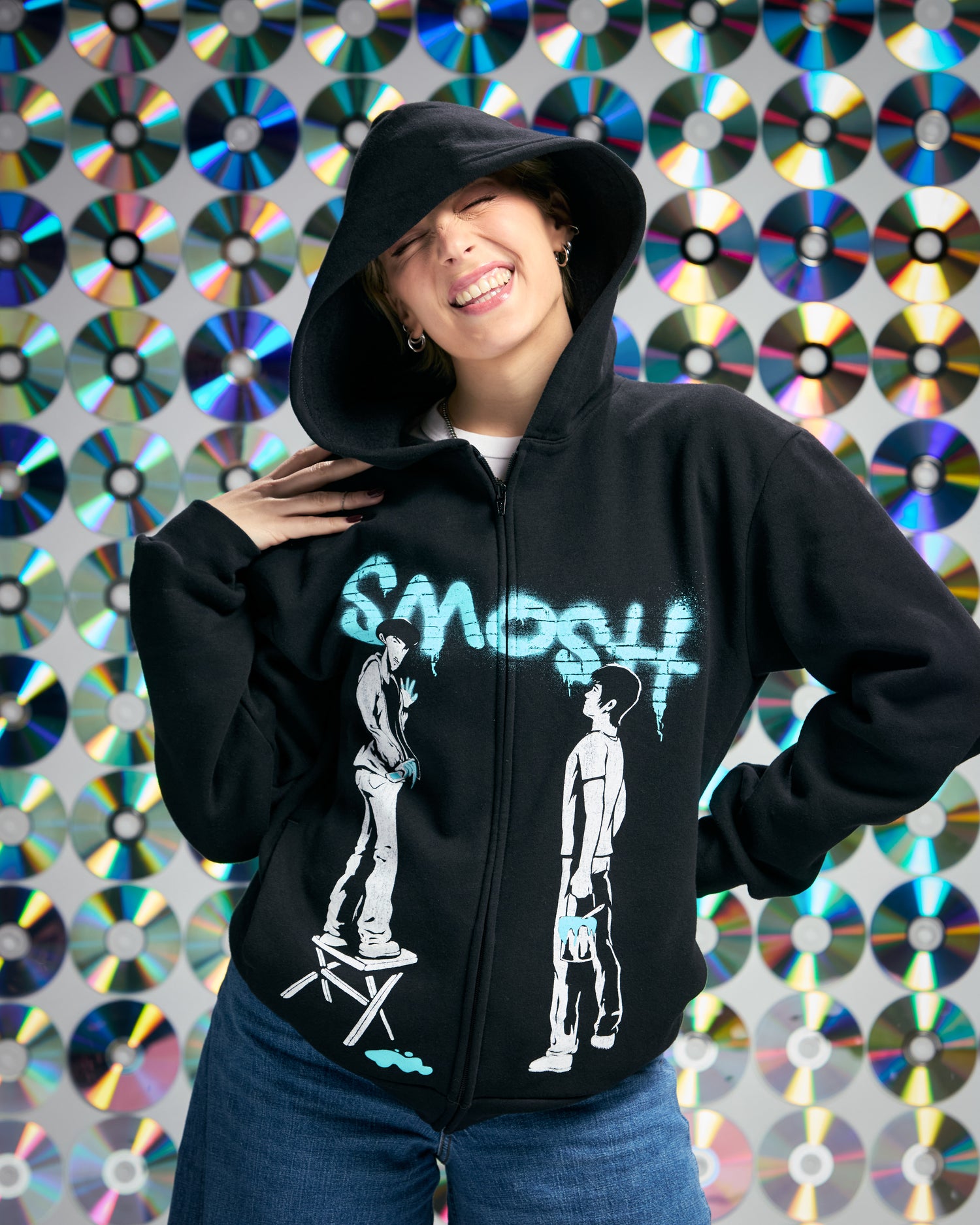 Person wearing a black hoodie with graphic design in front of a colorful CD wall.