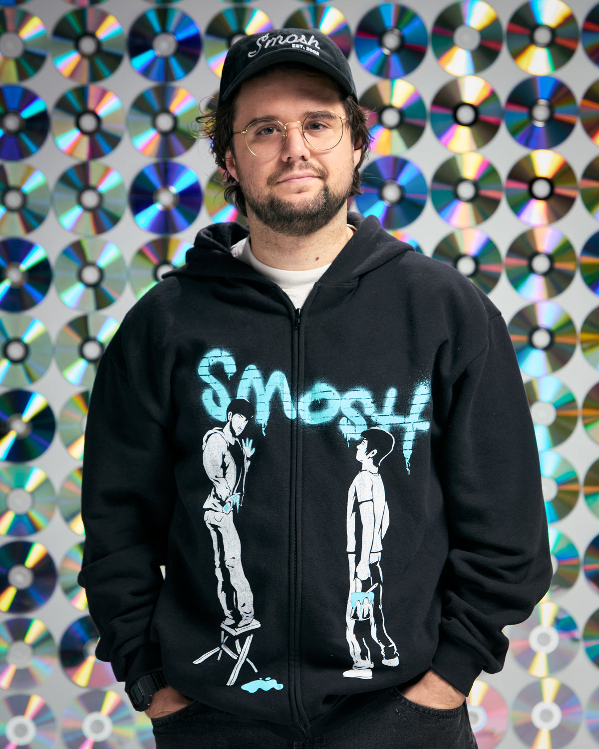 Person wearing a black hoodie with Smosh graphic and text, standing against a colorful CD disc background.