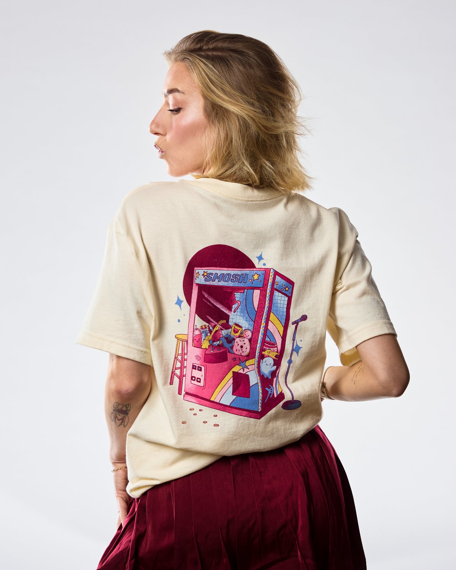 Woman wearing a cream t-shirt with 'SMOSH' text and stars on a white background back of shirt has a claw machine 